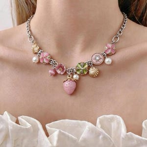 May include: Silver-toned necklace featuring pink flower, strawberry, and seashell charms. The necklace includes faux pearls and gold-toned accents. The chain is a linked design, and the necklace is worn around the neck.