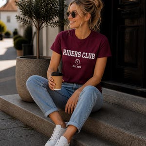 Könnte beinhalten: Ein kastanienbraunes Kurzarm-T-Shirt mit dem weißen Schriftzug RIDERS CLUB, einem kleinen weißen Pferdekopf-Symbol und dem Text EST. 2025. Das Oberteil wird mit einer hellblauen Jeans und weißen Turnschuhen zu einem lässigen Outfit kombiniert.