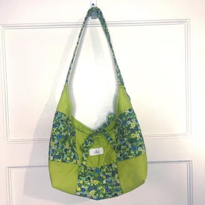 May include: A lime green hobo bag featuring a patchwork design with floral fabric panels in shades of blue, yellow, and green. The bag includes a long shoulder strap and a small white rectangular label with black text centred on the front.