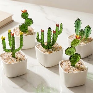 May include: Five small, decorative cactus plants in white square pots. The cacti are made of green glass with colorful flower accents. The pots are filled with small, light-colored pebbles, and the scene is set on a white surface.