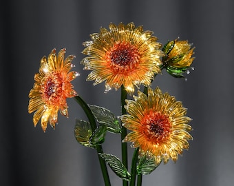 Puede incluir: Una escultura decorativa de cristal que presenta tres girasoles en flor y un capullo. Las flores tienen pétalos translúcidos amarillos y naranjas con centros marrones texturizados, sobre tallos y hojas de cristal verde.