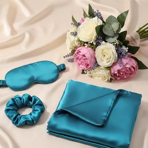 May include: A teal-colored sleep set including an eye mask, scrunchie, and a folded satin sheet. A bouquet of white and pink roses, lavender, and greenery sits nearby. The items are arranged on a cream-colored satin surface.