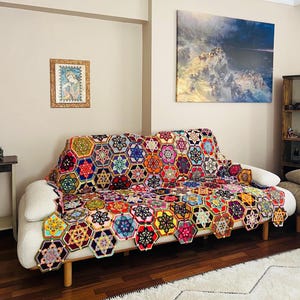 May include: A colorful, handmade crochet blanket draped over a white sofa. The blanket features a geometric pattern of hexagonal shapes in various colors, including red, blue, yellow, and green. A framed artwork hangs on the wall behind the sofa.