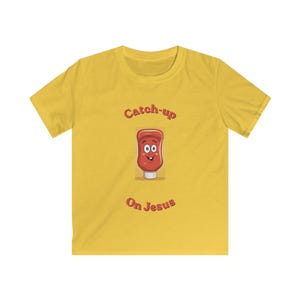 May include: Yellow t-shirt with the text "Catch-up On Jesus" in a playful font. The design features a cartoon ketchup bottle with a smiling face. A fun and lighthearted graphic tee.