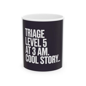 May include: A white ceramic mug with a black exterior. The mug features the text "TRIAGE LEVEL 5 AT 3 AM. COOL STORY." in white, bold, uppercase letters. The mug has a white interior and a standard handle.