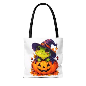 Puede incluir: Un bolso tote blanco con asas negras que presenta un gráfico de una rana verde con un sombrero de bruja morado, sentada dentro de una calabaza naranja tallada rodeada de hojas de otoño sobre un fondo blanco.