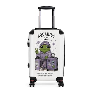 May include: White rolling suitcase with a cartoon frog wearing a purple hoodie and beanie. The word "Aquarius" is at the top. The frog is giving a peace sign. A small suitcase and water bottle are next to the frog. Text at the bottom reads "Different by Nature, Legend by Choice."
