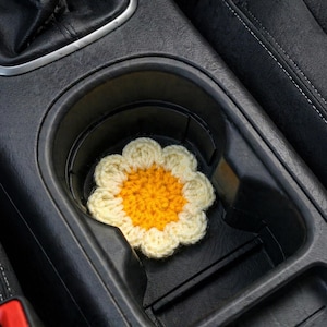 Può includere: Un sottobicchiere per auto all'uncinetto a forma di margherita, con centro giallo e petali color crema, si trova all'interno del portabicchieri di un'auto. Il sottobicchiere aggiunge un tocco decorativo agli interni dell'auto.