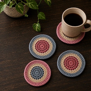 Puede incluir: Cuatro posavasos de ganchillo hechos a mano en tonos rosa, beige y azul, dispuestos sobre una superficie de madera oscura. Un posavasos sostiene una taza beige llena de café. Una planta en maceta con hojas verdes está en el fondo.