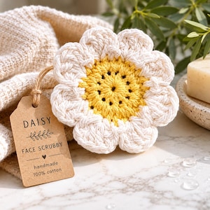 May include: A handmade daisy face scrubby made of 100% cotton. The scrubby is white with a yellow center and is shaped like a flower. A brown tag with the word "DAISY" is attached to the scrubby.