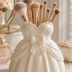 May include: A white ceramic makeup brush holder shaped like a ballgown with a central bow at the waist and beaded trim along the neckline and skirt. The holder contains a collection of various makeup brushes with rose gold ferrules and soft bristles.