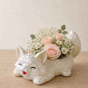 Op de afbeelding: Een parelmoer witte keramische plantenbak in de vorm van een kat met een textuur van vacht, puntige oren en een lachend gezicht met een rode mond. De plantenbak is gevuld met lichtroze rozen, kleine madeliefjes en gipskruid.
