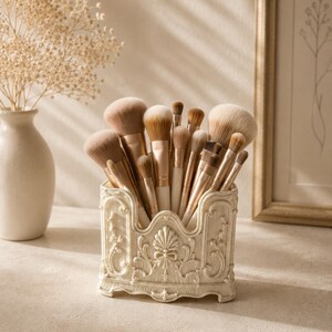May include: A collection of makeup brushes with light brown bristles and metallic rose gold ferrules, housed in an ornate, cream-coloured rectangular resin holder featuring intricate relief carvings of floral motifs and a central shell design.