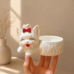 Op de afbeelding: Een witte, hondvormige plantenbak met een rode strik op zijn hoofd. De plantenbak heeft een lachend gezicht en een kleine, open kom voor het vasthouden van planten of kleine voorwerpen. De vacht van de hond is getextureerd en de plantenbak wordt in een hand gehouden.