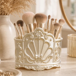 Op de afbeelding: Een crèmekleurige make-up kwastenhouder met een sierlijk, verhoogd schelpvormig reliëf aan de voorzijde. De houder is voorzien van ingewikkeld krulwerk, een klein strikdetail en een parelmoer afwerking, gevuld met make-up kwasten met gouden hulzen.