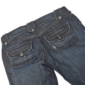 Könnte beinhalten: Dunkelblaue Jeans mit zwei Gesäßtaschen und Knopfdetails. Die Jeans haben ein klassisches Fünf-Taschen-Design und einen leicht abgenutzten Look. Die Taschen haben Knopfverschlüsse.
