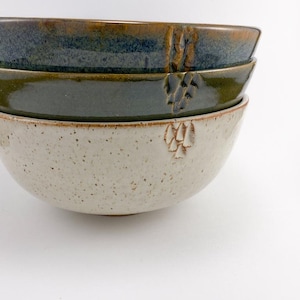 May include: A stack of three ceramic bowls. The top bowl is dark blue and brown, the middle bowl is olive green, and the bottom bowl is speckled off-white. Each bowl has a carved grape cluster design.