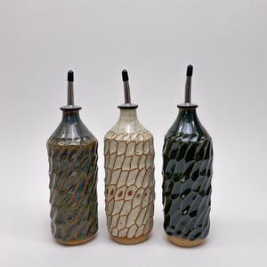 May include: Three ceramic oil dispensers with textured, faceted surfaces. The left bottle is blue-green, the centre is cream with brown accents, and the right is dark green. Each features a narrow neck and a silver metal pour spout with a black cap.
