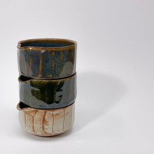 May include: A stack of three ceramic pouring bowls with a small spout on each rim. The top bowl is a deep blue with brown streaks, the middle bowl is a solid dark olive green, and the bottom bowl is a textured off-white with vertical brown grooves.