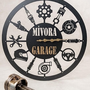 Peut inclure: Horloge de garage en métal noir avec des silhouettes de pièces automobiles sur le pourtour. Le cadran de l'horloge porte les mots "Mivora Garage" et des aiguilles dorées. Un piston, un ressort et des clés sont au premier plan.