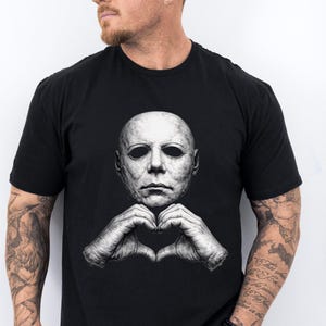 May include: A black short-sleeved t-shirt featuring a graphic print of a pale, textured human-like mask with dark eye sockets. Below the mask, two hands are positioned to form a heart shape. The design is rendered in shades of gray and white.
