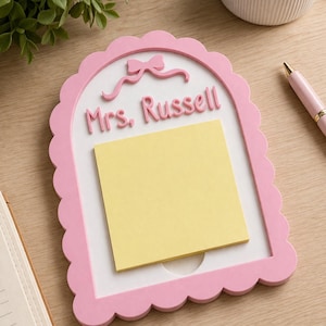 May include: A pink and white memo board with a scalloped edge, featuring the name "Mrs. Russell" in raised letters and a bow design. A yellow sticky note pad is attached to the board. A pink pen is visible in the background.
