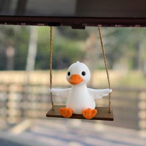 May include: A white duck figurine with orange feet and beak is suspended from a car's rearview mirror. The duck is seated on a small wooden swing, adding a whimsical touch to the vehicle's interior. The background is blurred.