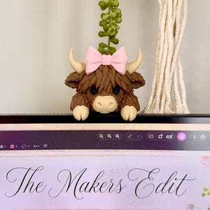 May include: A brown Highland cow monitor buddy with a pink bow, sitting atop a computer monitor. The monitor displays the text "The Makers Edit" and "Highland Cow Monitor Buddy" with floral accents.