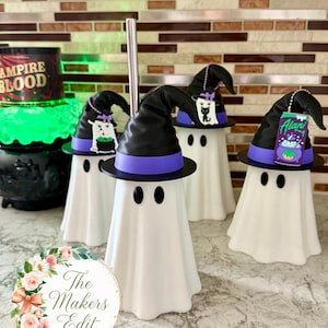 May include: Four white ghost-shaped tumblers, each wearing a black witch hat with a purple band. One tumbler features a silver metal straw. The hats are decorated with small charms, including a cat, a cauldron, and a purple Alani drink can.