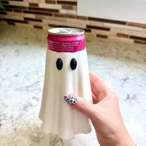 Può includere: Un manicotto refrigerante per lattine stampato in 3D a forma di fantasma bianco con due occhi ovali neri. Il manicotto è inserito su una lattina rosa Red Bull The Peach Edition, che mostra testo tra cui The Taste of White Peach e The Wings of Red Bull.