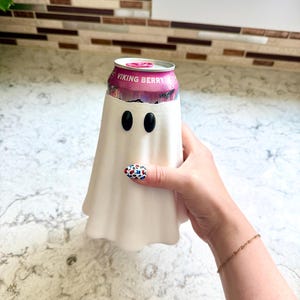 May include: A white, ghost-shaped beverage can cooler featuring two black oval eyes. The cooler is designed to fit around a standard aluminium can, which is visible at the top with a pink label that reads VIKING BERRY in white capital letters.