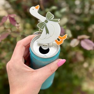 Puede incluir: Un abridor de latas con forma de ganso blanco con patas naranjas y un lazo verde está sujeto a una lata turquesa. El abridor de latas se sostiene en una mano con esmalte de uñas rosa. El fondo es follaje verde y rosa borroso.