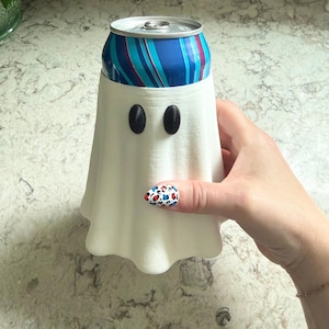 May include: A white, ghost-shaped beverage can cooler sleeve featuring two small, oval black eyes. The sleeve is designed to fit around a standard metal drink can, which has a blue, red, and white striped pattern visible at the top opening.