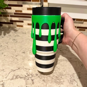 Könnte beinhalten: Ein hoher Becher mit einem horizontalen schwarz-weiß gestreiften Muster und einem leuchtend grünen, 3D-gedruckten Schleim-Tropfen-Überzug. Der Becher hat einen schwarzen Deckel, einen Metallstrohhalm und einen seitlich angebrachten schwarzen Griff.