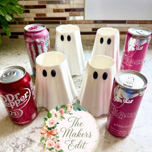 May include: Four white 3D-printed ghost-shaped cup sleeves with black oval eyes, arranged among three beverage cans: a red Dr. Pepper can, a pink Monster Viking Berry can, and a pink Red Bull The Peach Edition can. A circular logo reads The Makers Edit.