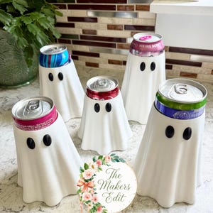 Pode incluir: Cinco capas para latas de bebida em forma de fantasma branco, cada uma com dois olhos ovais pretos. As capas estão colocadas sobre várias latas de refrigerante de alumínio coloridas. Um logótipo circular com detalhes florais e o texto The Makers Edit está centrado.