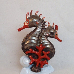 May include: A porcelain figurine of two seahorses with red coral and a white shell base. The seahorses are silver and have red eyes.