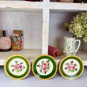 May include: Three decorative plates with a green border and a gold rim. Each plate features a floral design with pink roses and green leaves. The plates are arranged on a white surface.