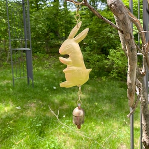 May include: A gold-colored rabbit wind chime with a bell hanging from a chain. The chime is hanging from a tree branch.