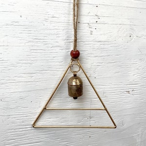 Könnte beinhalten: Ein goldener Dreiecks-Windspiel mit einer Glocke, die in der Mitte hängt. Das Windspiel ist an einer braunen Bindfadenkordel mit einer roten Holzperle befestigt.