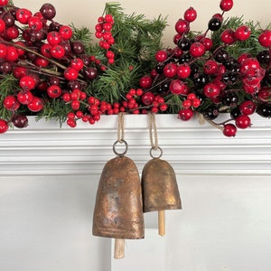 May include: Two antique-looking copper bells with wooden clappers hanging from a white mantelpiece decorated with a garland of red and black berries.