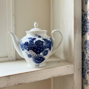 May include: A white porcelain teapot with a blue floral design sits on a white shelf. The teapot has a curved spout and handle, and a small lid. Blue and white toile curtains are visible in the background.