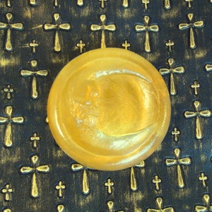 Peut inclure: Un savon rond, jaune doré et translucide, avec un motif de croissant de lune. Le savon est posé sur une surface noire avec un motif répétitif de croix dorées.