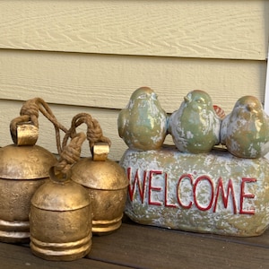 May include: Three gold bells with a rope attached, hanging from a welcome sign with three birds perched on top. The sign is made of a light green material with the word "WELCOME" written in red.