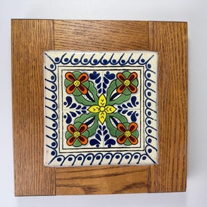 May include: Square decorative tile with a floral design in a wooden frame. The tile features a central yellow flower, surrounded by green leaves and orange and red flowers, with a blue and white border. The frame is made of light brown wood.