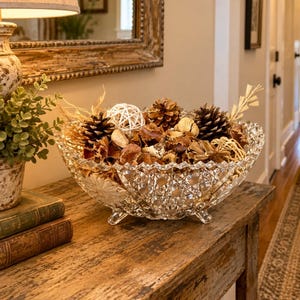May include: A clear, footed crystal bowl with a diamond-cut pattern filled with a collection of natural decorative items including brown pinecones, dried seed pods, and small white woven spheres, creating a textured centerpiece arrangement.
