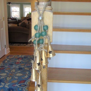 May include: A wooden stair banister with a decorative rope and green glass beads hanging from it. The beads are attached to a string of gold bells with star cutouts.