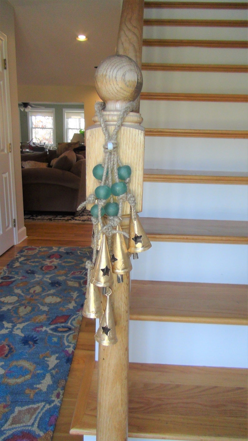 May include: A wooden stair banister with a decorative rope and green glass beads hanging from it. The beads are attached to a string of gold bells with star cutouts.