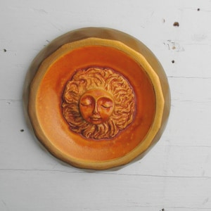May include: A round, orange ceramic wall plaque with a raised, sculpted face of a woman with closed eyes. The face is surrounded by a halo of hair and the plaque has a gold rim.
