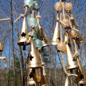May include: A set of gold-colored bells hanging from a rope with blue and pink glass beads. The bells are cone-shaped with a star cut-out and are hanging in a forest setting.